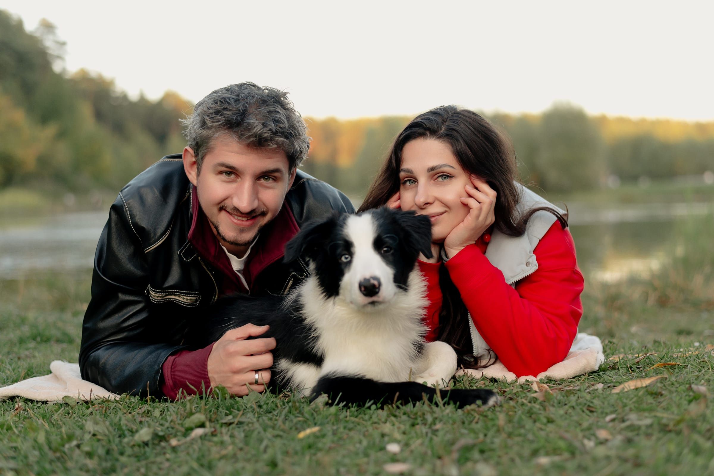 Vlad and Anna lying on the grass by the water, with Nox, their black-and-white border collie, between them