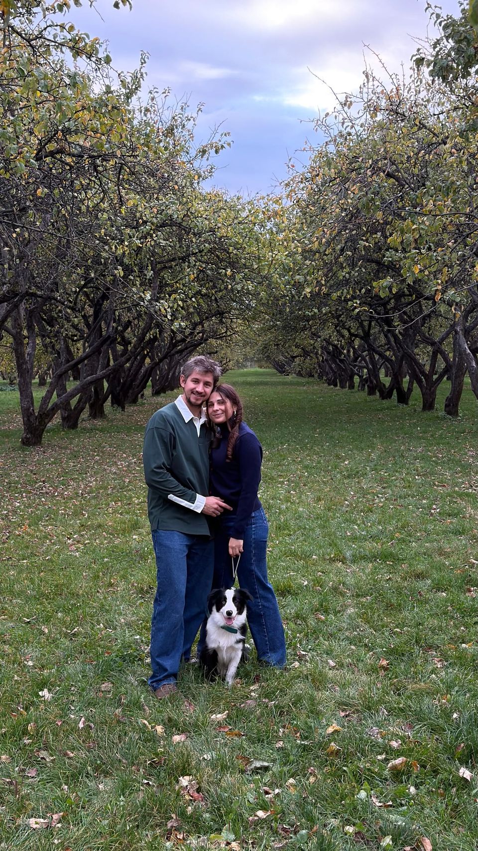 Vlad and Anna hugging in an autumn orchard, with Nox sitting at their feet